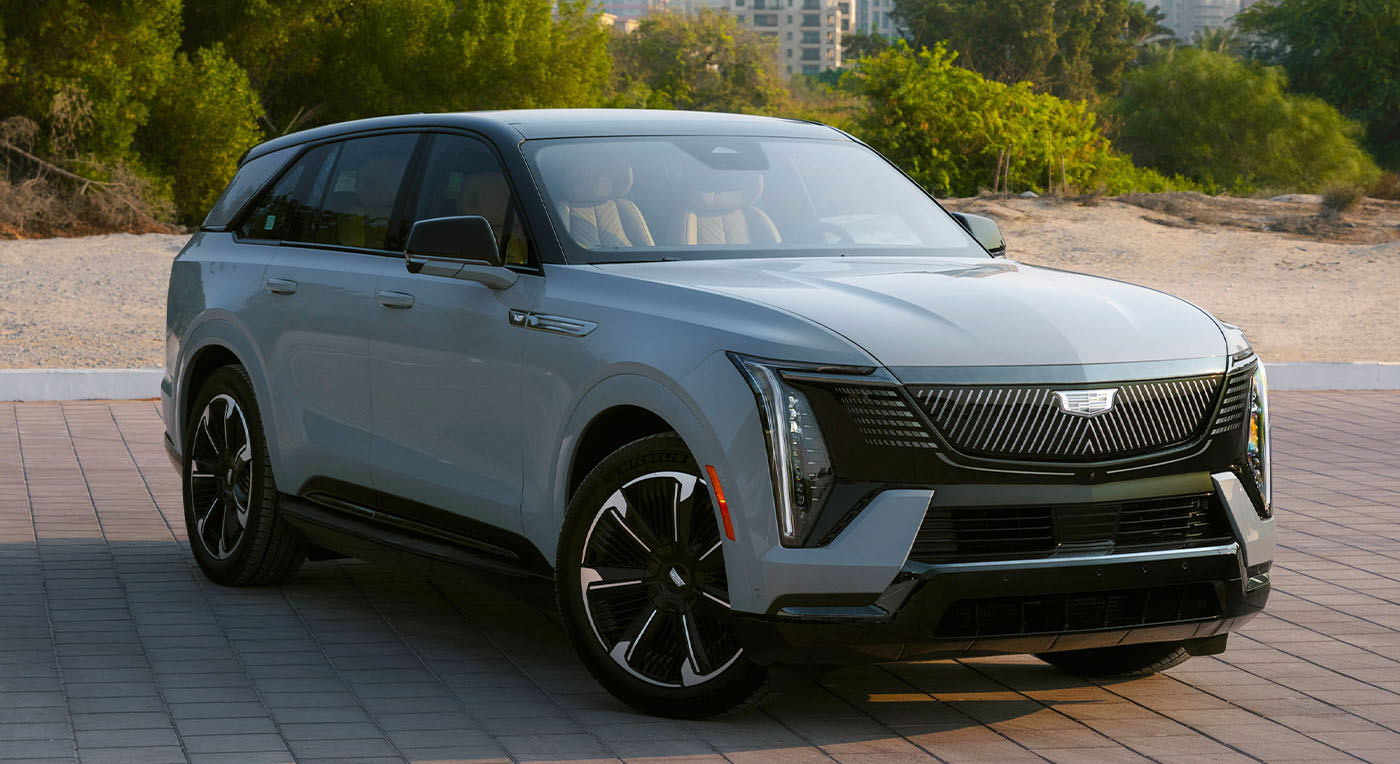 A sleek Cadillac Escalade IQL light gray SUV with a bold front grille and sharp headlights parked on a paved surface, framed by lush greenery and distant cityscape.